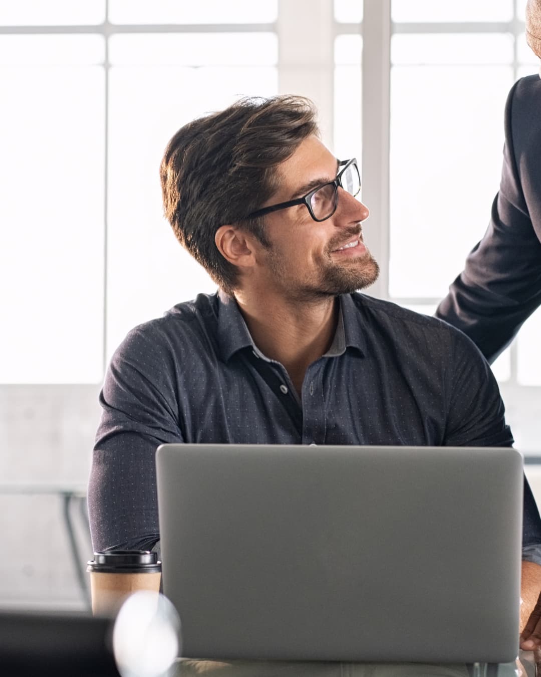 A man working at a laptop, looking up to talk with a colleague
